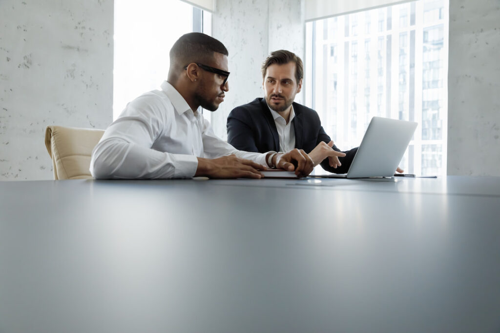 two businessmen sit at a large desk while discussing important work on the laptop in front of them