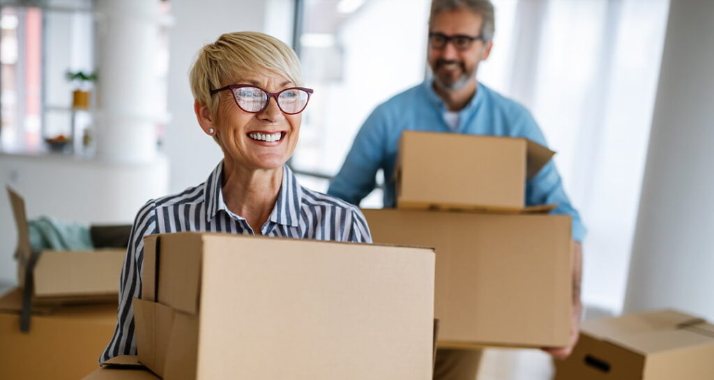 Older woman holding a cardboard moving box smiles as she walks with it through a room that is being packed up.