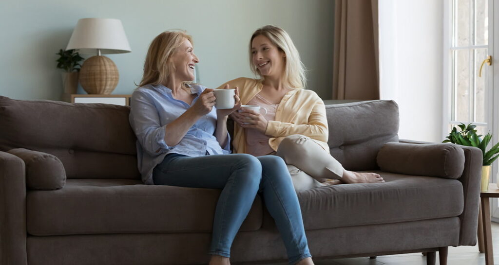 Two women sitting on a couch at home, smiling and enjoying coffee together in a warm, relaxed setting near a window.