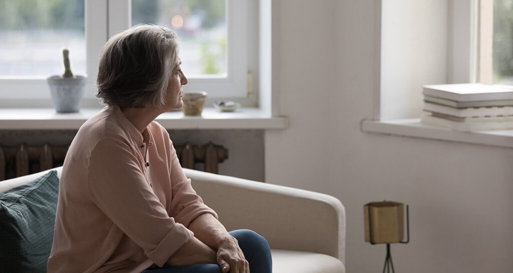 Older woman with short gray hair sitting on a couch, looking thoughtfully out a window in a softly lit room. A potted cactus, books, and other small items are visible on the windowsills.